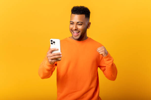 Happy satisfied man holding smartphone and smiling making yes gesture, celebrating online lottery or giveaway victory. Indoor studio shot isolated on orange stock photo