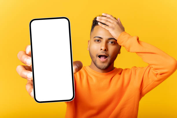 istock Surprised smiling man freelancer showing and holding smartphone with empty screen, amazed with new mobile app, received offer. Indoor studio shot isolated on yellow background 1468889235