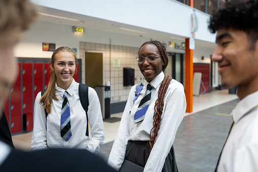An over-the-shoulder view of students stood outside of their classroom in the high school that they go to in the North East of England. They are dressed in their school uniform and chatting as they wait.