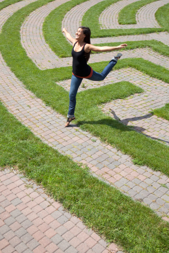 Beautiful young Asian woman playfully jumps over the grass boundary of a park labyrinth. Vertical shot.