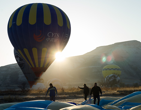 Goreme; Turkey - November 12; 2017: The crew of assistants folding hot air balloon that has landed after the flight at spectacular Cappadocia. Sun and silhouettes of hot air balloons on background