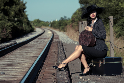 A young woman in old fashioned dress sits on her traveling trunk waiting for the train to come. A small suitcase sits in her lap as she waits