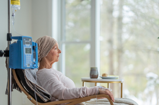 A senior woman sits in the comfort of her own home as she receives her chemotherapy treatment.  She is dressed comfortably and wearing a headscarf to keep her warm.