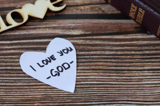 I love you-God, handwritten text on heart-shaped note and closed holy bible book on a wooden table. A closeup. Christian concept of love and hope in Jesus Christ.