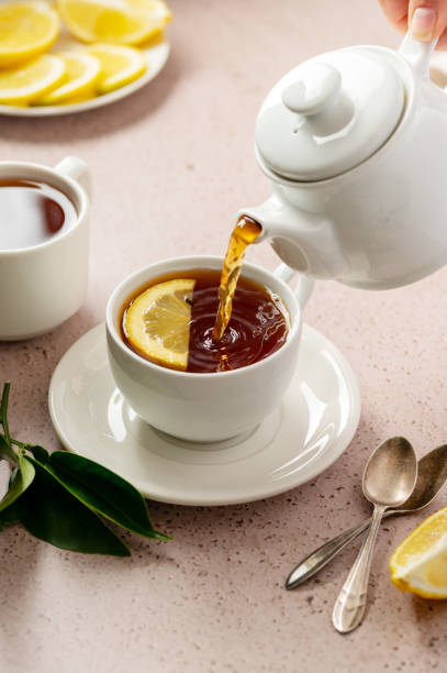 istock Pouring tea from tea pot, white ceramic cup of black tea and lemon slice. Vertical image. 1468917077