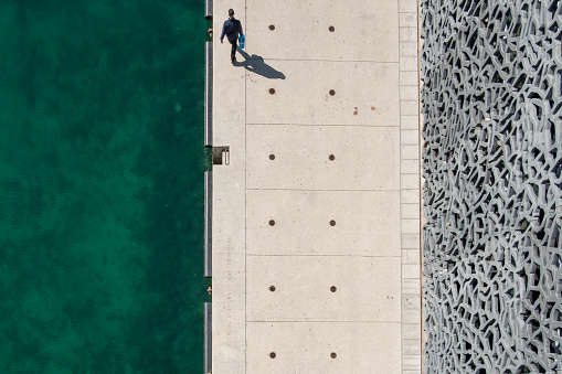 Top view of a human walking on a pier between the sea and a building