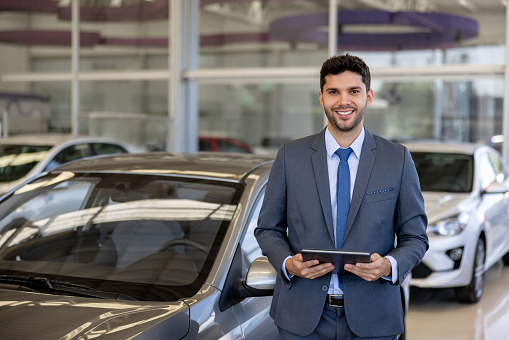 Happy Latin American salesman working at a car dealership and using a tablet computer while looking at the camera smiling