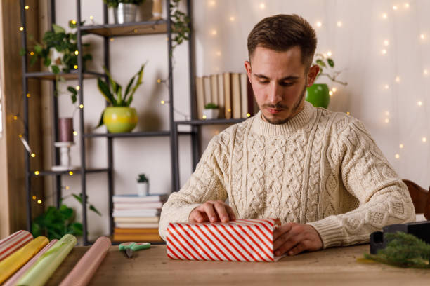 jeune homme rusé emballant soigneusement une boîte cadeau - christmas office gift people photos et images de collection