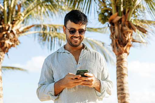 Smiling man using smartphone on tropical beach under shade of palm trees