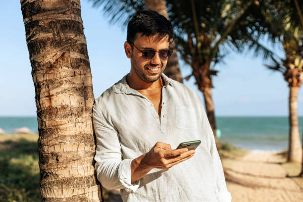 istock Smiling man using smartphone on tropical beach under shade of palm trees 1468925552