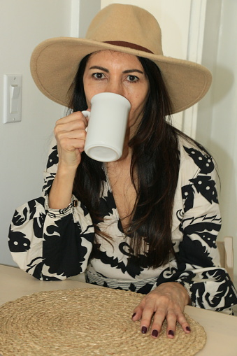 A Chilean woman drinking coffee from a large mug. She is wearing long brow straight hair, a hat and a black and white pattern dress.