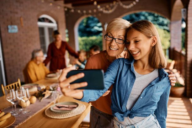 Happy teenage girl and her grandmother taking selfie during family gathering on a patio. Happy senior woman and her teenage granddaughter having fun while taking selfie on a terrace. Their family is in the background. teenage girl with grandmother stock pictures, royalty-free photos & images