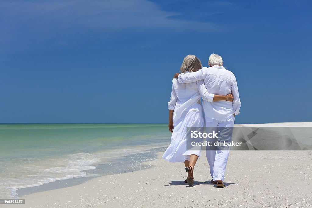 Rear view of senior couple walking on beach A senior couple with their arms wrapped around one another walks along a sandy tropical beach.  They are both wearing all white, with the exception of their footwear. In the background, a clear blue sky provides contrast to the all-white attire.  The tide flows on the shore, nearly touching the couple's feet. Senior Adult Stock Photo