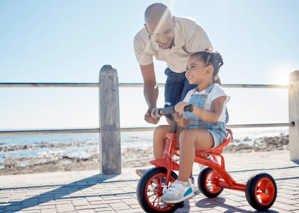 niño, papá y aprendiendo a andar en bicicleta en el paseo marítimo, el sol y el verano al aire libre para divertirse, desarrollarse y jugar. padre enseñando ciclismo a niña feliz en bicicleta en la acera del océano en la naturaleza - beach family boardwalk footpath fotografías e imágenes de stock