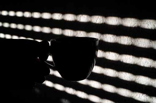 Silhouette of a coffee cup in the morning with light streaming through window shutters on the wall