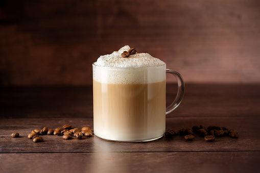Glass cup of layered coffee drink with milk foam, coffee beans over wooden background, copy space.