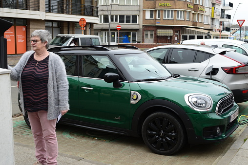 Blankenberge, West-Flanders, Belgium - February 21, 2023: woman at a parking payment terminal in foreground of Mini-Cooper next to a electric charging station square Leopold 3 in Blankenberge