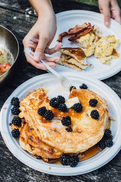 stack of pancakes with maple syrup, blackberries, butter on paper plates on outdoor picnic table