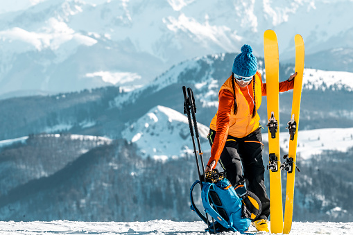 A fully and colorfully equipped woman with proper skiing clothing, ski boots, cap, sunglasses  and a backpack seen standing and holding skis after finishing her ski touring adventure during one sunny winter day.