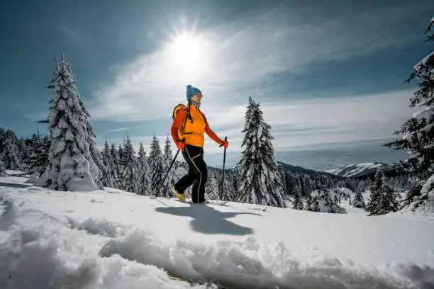 A colorfully dressed woman with all proper winter equipment like ski jacket, pants, woolen cap, skis, poles, ski boots, backpack and sunglasses seen ski touring in a beautiful nature of the mountain covered with snow during her winter vacation.