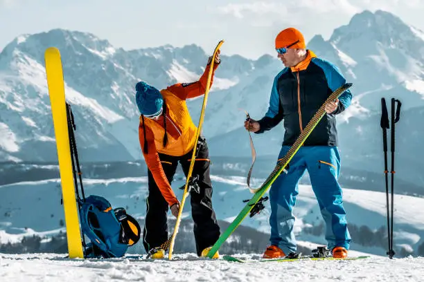 A fully equipped couple with proper skiing clothing, ski boots, cap, sunglasses  and a backpack seen holding skis and taking off the skins after finishing their ski touring adventure during one sunny winter day with mountains in the background.