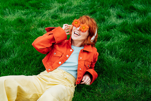 Happy smiling redhead woman with freckles in red orange gerbera Flower glasses lying on green grass and listening to music in wireless headset. Positive Emotions. Fashion. Spring, summer mood