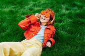 Happy smiling redhead woman with freckles in red orange gerbera Flower glasses lying on green grass and listening to music in wireless headset. Positive Emotions. Fashion. Spring, summer mood.