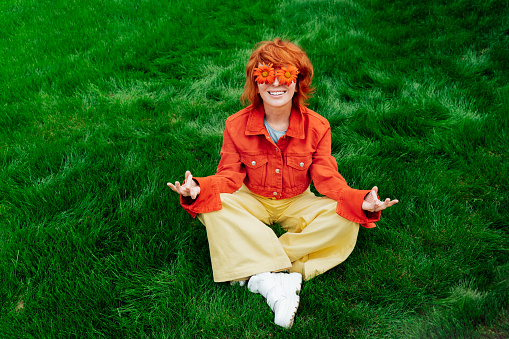 Relaxed Happy smiling redhead Woman with freckles and red orange gerbera Flower sunglasses sitting in meditation pose on green grass and enjoy the moment. Positivity vibe Emotion. Spring, summer mood.