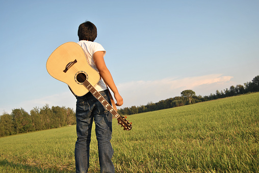 A teenager stands in an open field with his guitar on his back. Shallow DOF.