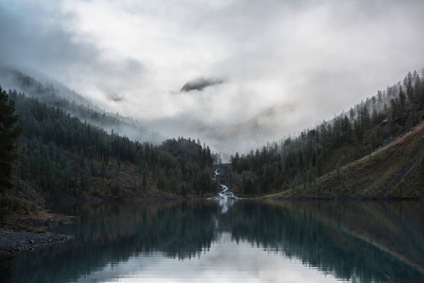 istock Mountain creek flows from forest hills into glacial lake. Tranquil scenery with rocks in clearance of mysterious fog. Small river and coniferous trees reflected in calm alpine lake in early morning. 1469001497