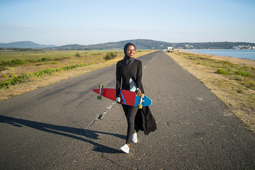 A young African-American woman is holding a skatebaordwith one hand, wearing modest clothing and smiling into the distance as she walks along an empty highway under a clear blue sky