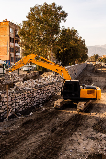 A digger working during the daytime. The machine works in a stream bed, digging the ground. Streets are visible. He works under a bridge.
