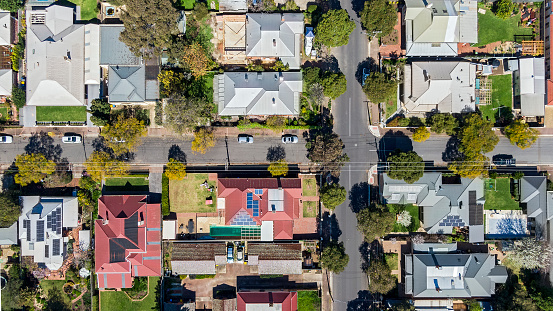 Aerial view directly above established houses in leafy Adelaide suburb: variety of architectural styles and roofing materials, cars parked in street and driveways, solar panels on some houses, treelined streets with overhead powerlines.