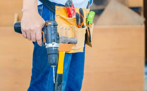 Photo of Handyman manual worker in tools belt and holding drill in his hands, Carpenter working with equipment in wood workshop, man doing woodwork in carpentry shop