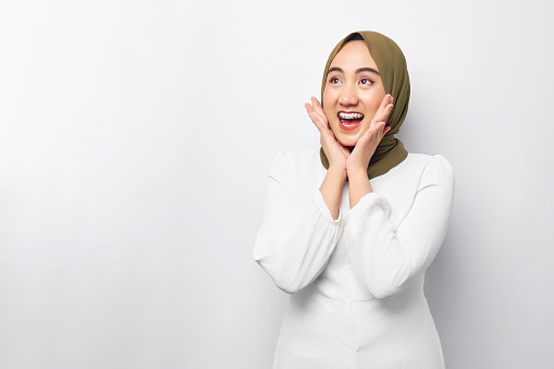 Excited young Asian Muslim woman 20s wearing hijab looking at copy space and raising hands with open mouth, reacting to sale offer isolated on white background. People religious lifestyle concept