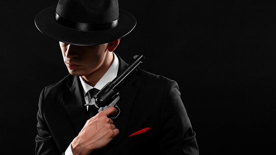 Stylish portrait of gangster from 1940s with a gun. Man in a black suit and hat with a gun over dark background.