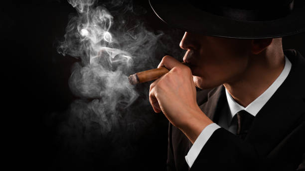 Retro style photo of a shaded detective in a black suit and hat holding and smoking cigar over black background. Retro style close-up photo of a shaded detective in a black suit and hat holding and smoking cigar over black background. cigar  stock pictures, royalty-free photos & images