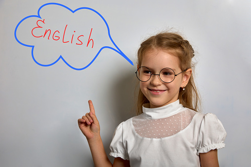 Joyful girl in white blouse is standing at blackboard in English class, pointing finger at blackboard with inscription English. Schoolgirl in classroom smiles while standing at blackboard in classroom