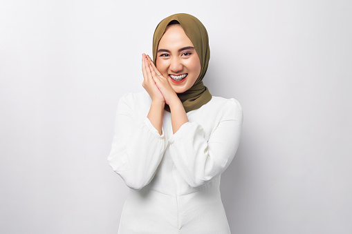 Glad satisfied young beautiful Asian Muslim woman 20s wearing hijab touching her cheek and looking at the camera isolated on white background. People religious lifestyle concept