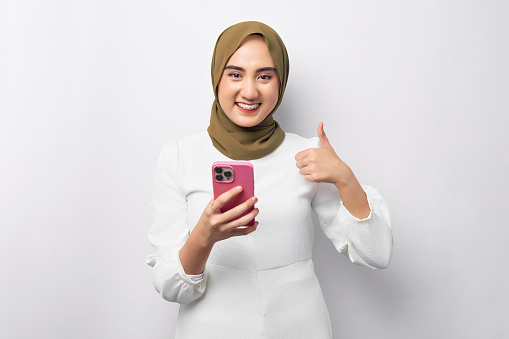 Beautiful smiling Asian Muslim woman wearing hijab Using mobile phone, typing sms message, showing thumb up isolated on white background studio portrait. People religious lifestyle concept