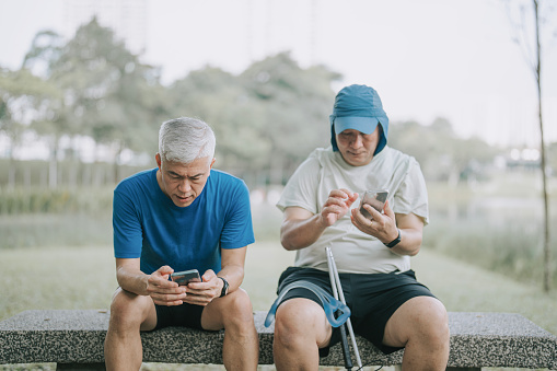 Asian Chinese visually impaired mature man sitting at park bench with smart phone together with his friend taking a rest