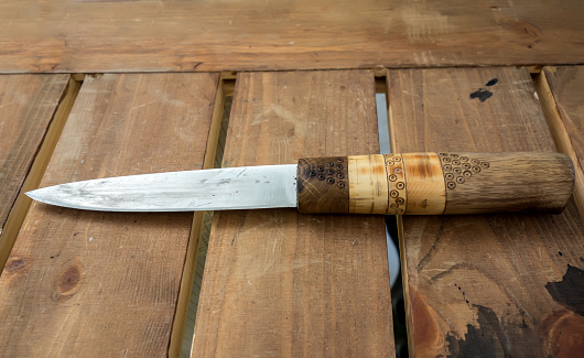 A sharp knife closeup with a wooden hand with a pattern lies on a wooden table. 
Knife for hiking or knife for entering the forest rustic background.