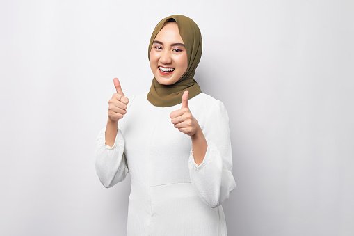 Beautiful smiling friendly young Arabian Asian Muslim woman wearing hijab showing thumb up gesture isolated on white background. People religious lifestyle concept