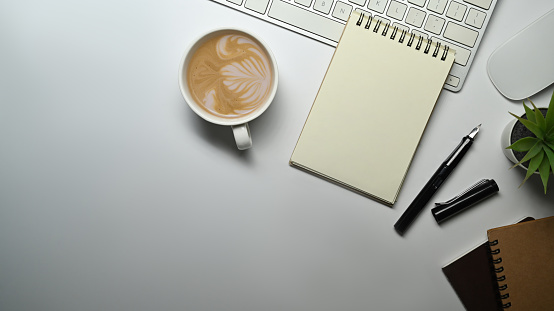 Top view of spiral notepad, pen, cup of coffee and keyboard on white office desk. Copy space for text.