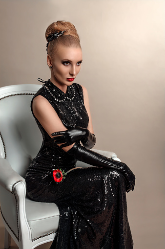 Блондинка в черном блестящем платье и черных перчатках сидит на белом стуле с красным цветком в руках. Шикарный студийный модный портрет