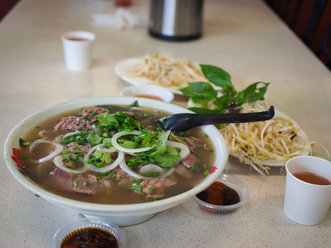 A bowl of delicious Vietnamese beef noodles soup known as 'Pho', garnished with chives and spring onions accompanied by a plat of bean sprouts, mint and chili sauces on a blurred background.