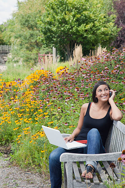 jovem mulher com computador portátil e cellphone em um banco de parque - using phone garden bench imagens e fotografias de stock