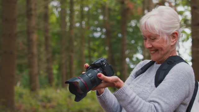 Retired active senior woman with DSLR camera taking photos on hike through woodland countryside walks into frame enjoying peace and beauty of nature- shot in slow motion