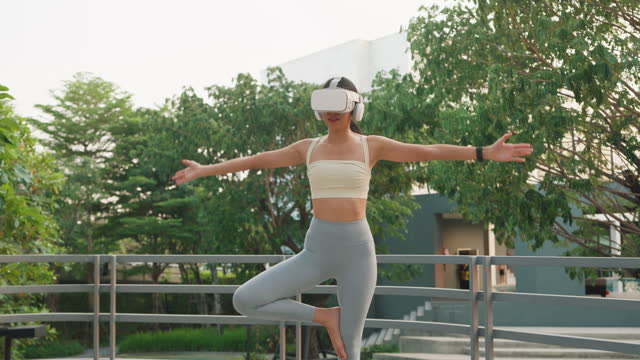 Young Asian athletic woman wearing virtual reality headset sportive top and leggings practicing yoga outdoors in city park. Diet and healthy weight loss.