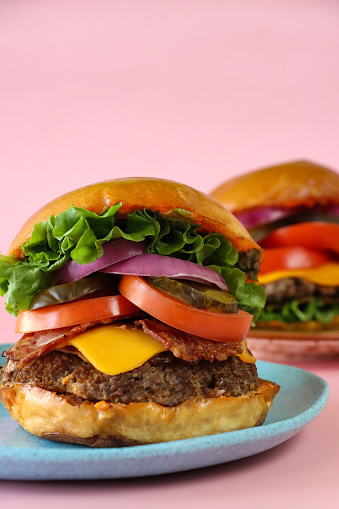 Stock photo showing a close-up view of the components of home-made gourmet burgers in burger buns consisting of meat patties, bacon rashers, melting processed cheese slices, lettuce leaves, sliced tomato, pickles and red onion slices, against a pink background.